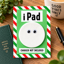 A joke iPad gift. A novelty card or sign styled on a wooden desk, featuring a white circular cutout with googly eyes and the text 'iPad' and 'CHARGER NOT INCLUDED' on a green background with red-and-white striped borders. Surrounding props include a green notebook, pen, mug, and plants.