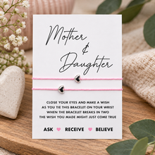 A Mother & Daughter bracelet gift set featuring a white card with instructions and a pink cord bracelet with two small silver heart charms, styled on a wooden surface with decorative props.