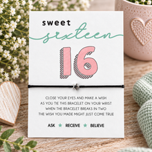 A sweet sixteen bracelet card displayed on a wooden surface with decorative flowers, a pink heart mug, and a knitted sweater. The card reads 'sweet sixteen 16' and includes instructions for tying the black cord bracelet with a star charm.