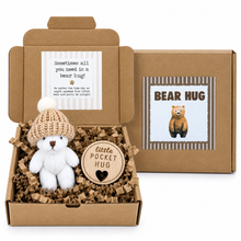 A teddy bear plush toy in a knitted cream hat displayed inside an open kraft gift box with shredded paper filler, alongside the closed box lid featuring a bear illustration and the text 'BEAR HUG'.