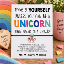 Unicorn wish string bracelet. A white card with colourful text reading 'Always be yourself unless you can be a unicorn then always be a unicorn,' tied with a black cotton string to a small rainbow charm bracelet.