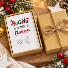 A pair of silver Christmas-themed stud earrings displayed in an open kraft gift box with a white insert that reads 'Believe in the Magic of Christmas.' 