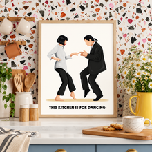 A framed art print featuring a retro illustration of a pulp fiction scene, with the text 'THIS KITCHEN IS FOR DANCING' at the bottom. The print is styled on a kitchen counter with a speckled terrazzo wall, wooden utensils, a yellow flower vase, and a coffee mug.
