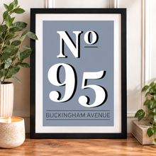 House number and road name print. A framed typographic art print reading “No 95 Buckingham Avenue” in large white serif numerals on a muted blue-gray background, displayed on a wooden surface with plants and a candle around it.