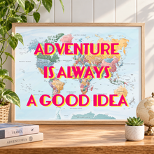 A framed world map poster with the phrase “ADVENTURE IS ALWAYS A GOOD IDEA” in bold pink letters, displayed on a wooden table with a plant, books, and a small succulent in a styled interior setting.