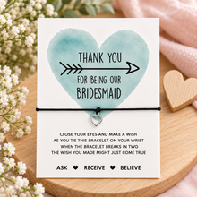 Bridesmaid wish string bracelet. A bridesmaid bracelet gift card. The card reads 'THANK YOU FOR BEING OUR BRIDESMAID' with a watercolour heart and arrow, and includes a small silver heart charm attached to a black cord bracelet.