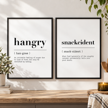 Two black-framed typography art prints hanging on a white wall above a wooden console table. The left print reads 'hangry' with a humorous dictionary-style definition, and the right print reads 'snackcident' with its own funny definition. Kitchen wall art.