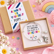Personalised unicorn charm bangle. A silver adjustable charm bracelet with a unicorn and letter P charms is presented in an open gift box next to its kraft paper packaging with a colourful 'Always Be Yourself unless you can be a unicorn. Then always be a unicorn. Unicorn gift 