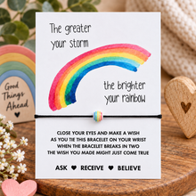 The greater your storm the brighter your rainbow wish string bracelet. A white card with a colourful watercolour rainbow and inspirational text, tied with a small striped bracelet across the middle, displayed on a wooden surface with decorative heart and rainbow props.