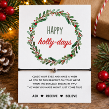 Happy holidays wish string bracelet. A festive white card with a green holly wreath and the words 'Happy holly-days' A black cord bracelet with a small silver star is tied across the centre of the card, with printed wish instructions below. Christmas gift 