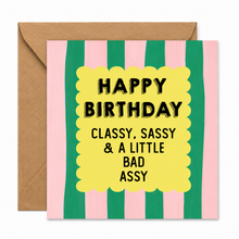 A square birthday card with pink and green vertical stripes, a yellow scalloped center panel, and black text reading 'HAPPY BIRTHDAY CLASSY, SASSY & A LITTLE BAD ASSY,' shown with a brown envelope behind it on a white background.