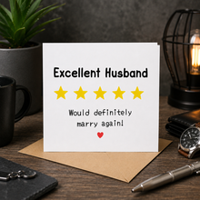 A white greeting card with the text 'Excellent Husband' and 'Would definitely marry again!' displayed on a desk with a kraft envelope, surrounded by a plant, mug, lamp, wallet, watch, and pen.