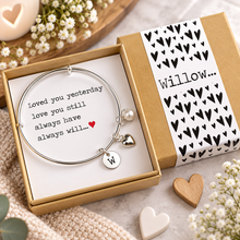 A silver bangle bracelet with a pearl charm, heart charm, and initial 'W' charm, presented in a kraft gift box with a printed message that reads. Loved you yesterday, love you still, always have always will. Gift for anniversary or valentines day