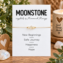 A gold beaded bracelet with clear crystal beads is displayed on a white card labeled 'MOONSTONE crystals by Mermaid Marys' with positive phrases printed below. The card is styled on a wooden surface with flowers, crystals, and a candle in the background.