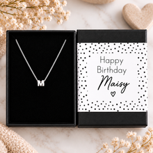 Personalised sterling silver letter necklace. A silver initial necklace with the letter 'M' displayed inside an open black gift box next to its closed lid, which features a 'Happy Birthday Maisy' message with black polka dots on a white background.