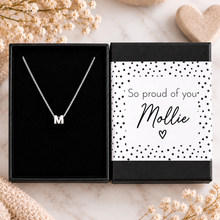 A silver initial necklace with the letter 'M' displayed inside an open black gift box next to its decorated packaging. The packaging features a white label with black polka dots and the text 'So proud of your Mollie'.