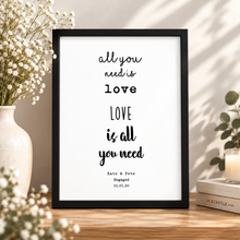 A black-framed typographic wall art print sits on a wooden surface in a styled interior scene. The print reads “all you need is love LOVE is all you need” personalised engagement print  