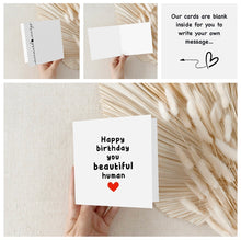 Blank greeting card with 'Happy birthday you beautiful human' text and heart, held against a textured beige background.